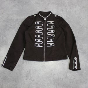 Könnte beinhalten: Dunkelbraune Jacke mit weißer Paspelierung und silbernen Knöpfen. Die Jacke hat einen hohen Kragen und einen Reißverschluss. Weiße Zierleisten verlaufen vorne und an den Manschetten. Jacke im Militärstil.