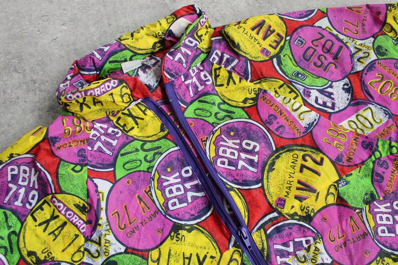 May include: A red, purple, yellow, and green patterned jacket with a zipper closure. The pattern features various state names and abbreviations, such as "Maryland", "Colorado", and "Washington", along with numbers and the word "PBK".