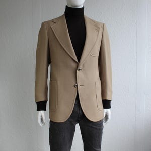 Może przedstawiać: Beżowa marynarka z dwoma guzikami i dwiema przednimi kieszeniami jest prezentowana na manekinie. Marynarka jest połączona z czarnym golfem i ciemnoszarymi dżinsami. Manekin ma białą głowę.