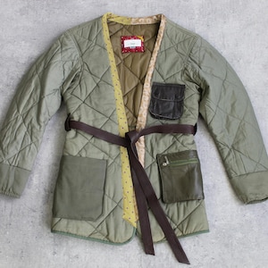 Könnte beinhalten: Gesteppte olivgrüne Jacke mit braunem Gürtel und Taschen. Die Jacke hat ein Rautenmuster und ein kontrastierendes gelb-braunes Innenfutter. Sie hat zwei Vordertaschen und eine dunkelbraune Ledertasche.