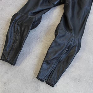 May include: Black leather motorcycle pants with knee padding and zippers at the ankles. The pants are lying on a gray surface. The leather appears worn and has visible stitching.