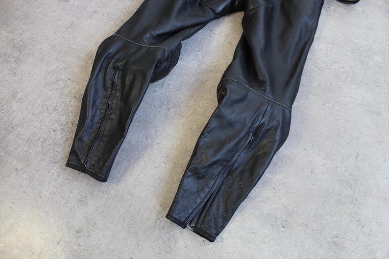 May include: Black leather motorcycle pants with knee padding and zippers at the ankles. The pants are lying on a gray surface. The leather appears worn and has visible stitching.