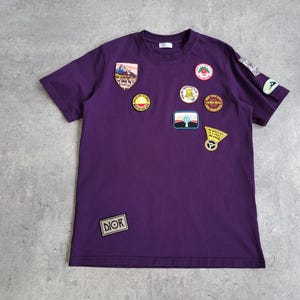 Peut inclure: Un t-shirt violet orné de divers écussons. Les écussons comprennent une scène de montagne, des motifs circulaires et le mot "DIOR". Le t-shirt a un col rond et des manches courtes.