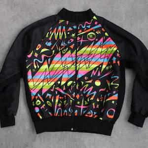 Op de afbeelding: Zwarte ritsjas met een kleurrijk abstract patroon op de voor- en achterkant. Het patroon bevat strepen, zigzaglijnen en geometrische vormen in roze, blauw, groen, geel en oranje.