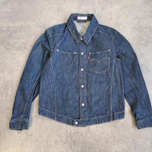 May include: A blue denim jacket with a button-down front, a collared neckline, and long sleeves. The jacket features a chest pocket with a red tag and white buttons. The jacket is made of a medium-wash denim fabric.
