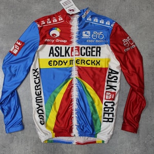 Pode incluir: Uma camisola de ciclismo de manga comprida branca, azul, vermelha e amarela. A camisola tem o texto "ASLK" e "Eddy Merckx" na frente e atrás. A camisola também tem uma faixa arco-íris na frente e atrás.