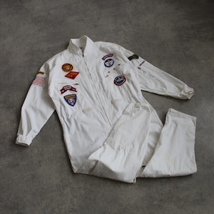 May include: White jumpsuit featuring several patches, including a US flag and military badges. The one-piece garment has a zip closure and long sleeves. The jumpsuit is displayed on a grey surface.