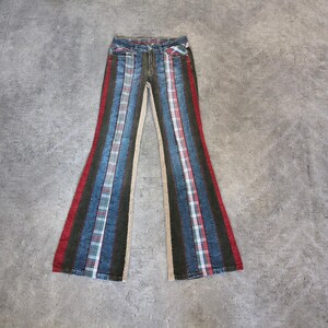 Op de afbeelding: Een uitlopende spijkerbroek met verticale panelen van blauwe denim, donkerbruine stof en rood, wit en blauw geruite stof. De broek heeft een standaard tailleband met knoopsluiting, voorzakken en een opvallend ontwerp met meerdere texturen.
