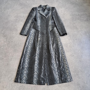 May include: A long coat with a reptile-like texture, featuring a notched collar, two front pockets, and a button closure. The coat is mainly grey with black and silver accents. It is laid flat on a grey surface.