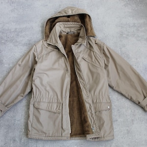 Może przedstawiać: Beżowa kurtka z kapturem, z kapturem podszytym futrem i zamkiem błyskawicznym. Kurtka ma dwie duże kieszenie z przodu i sznurek do regulacji w talii.