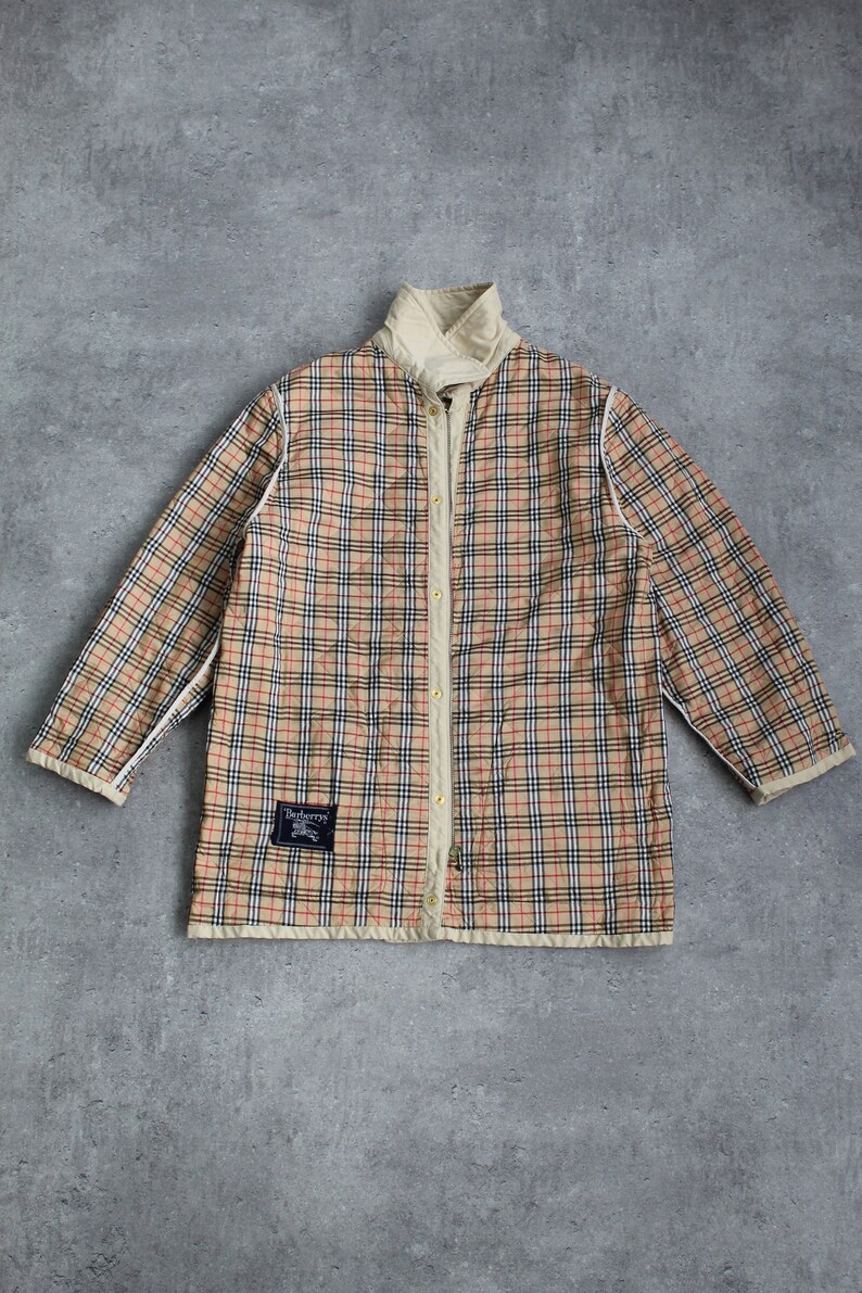 May include: A beige quilted jacket with a plaid pattern in black, red, and white. The jacket has a zipper closure and a stand-up collar. The label on the jacket reads "Burberrys".