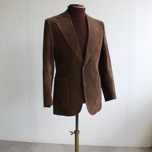 May include: A brown corduroy blazer with a notched lapel and two front pockets. The blazer is displayed on a vintage brass clothing rack. A brown turtleneck is visible underneath the blazer.