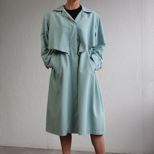 May include: A pale blue trench coat with a collared neckline and long sleeves. The coat has a button-down front, pockets, and a knee-length hem. The model is wearing a black top and beige heels.