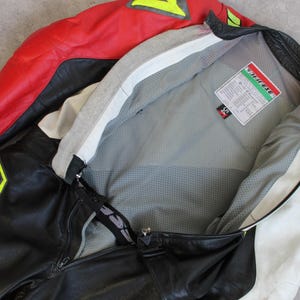 May include: A black, red, and white motorcycle jacket and pants set. The jacket has a red upper section and white accents, with a gray interior. The pants feature large yellow lettering. The set appears to be made of leather.