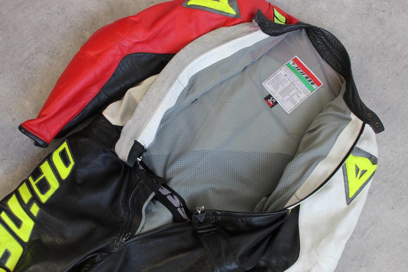 May include: A black, red, and white motorcycle jacket and pants set. The jacket has a red upper section and white accents, with a gray interior. The pants feature large yellow lettering. The set appears to be made of leather.