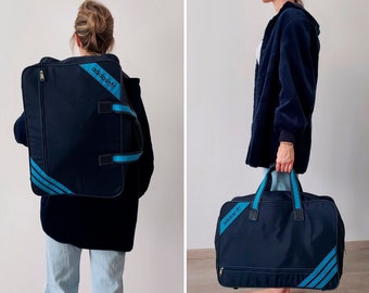 Pode incluir: Uma mala de viagem Adidas azul com listras azuis e brancas e o logotipo da Adidas. A mala tem um fecho de correr e uma pega superior.