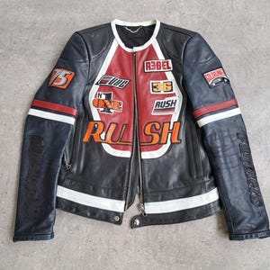 May include: Black leather motorcycle jacket with red and white detailing. Patches display the words "REBEL", "RUSH", and "ONE". It has a zip closure and white stripes on the sleeves. A fashionable outerwear item.
