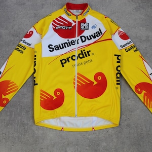 May include: Yellow and red cycling jersey with the text "Saunier Duval" and "prodir swiss pens". The jersey has a full-length zipper and long sleeves. Red and white accents are on the sleeves and shoulders. The jersey features a bird graphic.