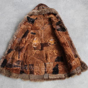 May include: A brown fur coat with a hood, featuring a patchwork design. The coat has embroidered details in gold and white, with a fur trim around the hood and edges. The coat is laid out on a gray surface.