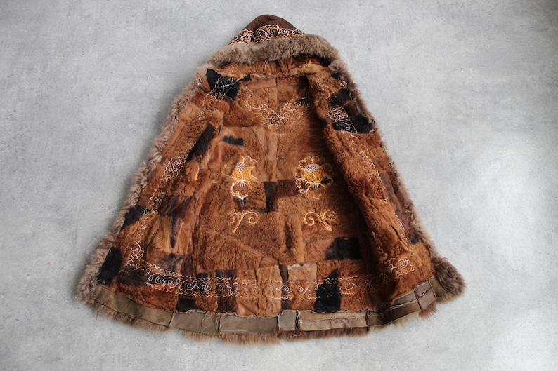 May include: A brown fur coat with a hood, featuring a patchwork design. The coat has embroidered details in gold and white, with a fur trim around the hood and edges. The coat is laid out on a gray surface.