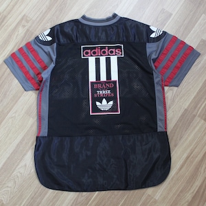 Op de afbeelding: Zwart en grijs Adidas-shirt met rode strepen en de tekst "adidas" en "Brand with Three Stripes" op de rug.