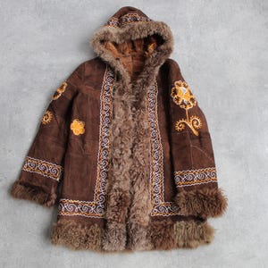 May include: A brown suede coat with a hood and fur trim. The coat features embroidered floral designs in yellow and white, and is lined with brown fur. The coat is laid flat on a gray surface.