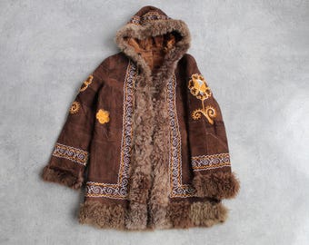 May include: A brown suede coat with a hood and fur trim. The coat features embroidered floral designs in yellow and white, and is lined with brown fur. The coat is laid flat on a grey surface.
