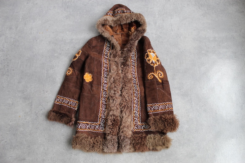 May include: A brown suede coat with a hood and fur trim. The coat features embroidered floral designs in yellow and white, and is lined with brown fur. The coat is laid flat on a gray surface.