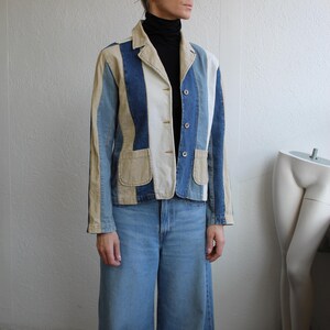Op de afbeelding: Een patchwork denim blazer met verticale strepen in licht- en middenblauw, beige en wit. De blazer heeft een kraag, twee voorzakken en drie knopen. Het model draagt de blazer met een lichtblauwe broek met wijde pijpen.
