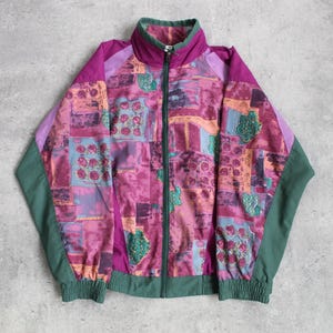 Op de afbeelding: Een vintage trainingsjack met een levendig patchwork-ontwerp. De jas heeft een volledige ritssluiting, een opstaande kraag en elastische manchetten en zoom. Het kleurenpalet omvat tinten roze, paars, groen en oranje, wat een gedurfde en retro esthetiek creëert.