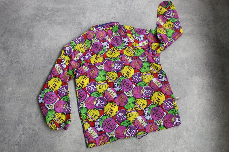 May include: A colorful button-down shirt with a pattern of various license plates. The plates are in shades of red, yellow, green, and purple, with black numbers and letters. The shirt has a long sleeve and a button-up front.