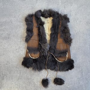 May include: A vintage brown leather vest with dark gray fur trim and accents. The vest has two front pockets and a cream-colored interior. Tassels hang from the bottom. A unique, rustic garment.