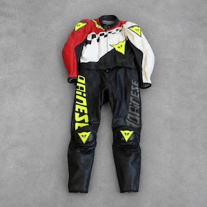 May include: A black and white motorcycle racing suit with red accents. The suit features yellow triangle logos and the word "DAINESE" in yellow and gray. The suit is made of leather and is designed for protection and speed.