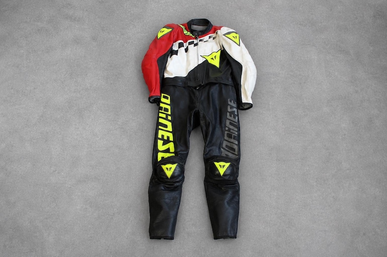 May include: A black and white motorcycle racing suit with red accents. The suit features yellow triangle logos and the word "DAINESE" in yellow and gray. The suit is made of leather and is designed for protection and speed.