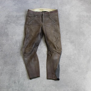Könnte beinhalten: Braune Lederhose mit einem abgenutzten, verwitterten Aussehen. Die Hose hat einen Knopf- und Reißverschluss, Gürtelschlaufen und Taschen. Die Beine verjüngen sich zu den Knöcheln. Die Hose wird auf einer grauen Oberfläche präsentiert.