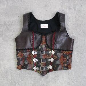Könnte beinhalten: Eine braune und schwarze Weste mit floralem Muster und silbernen Schnallen. Die Weste hat einen quadratischen Ausschnitt und ist mit schwarzer Spitze und roten Akzenten verziert. Die West hat ein Etikett mit Text.