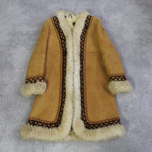 Puede incluir: Un abrigo color canela con interior y ribetes color crema. El abrigo presenta costuras decorativas marrones a lo largo de los bordes y puños. Los puños y el cuello están adornados con piel sintética color crema. Un abrigo de estilo vintage.