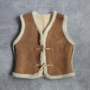 May include: A brown suede vest with cream-colored shearling lining and trim. The vest features two wooden toggle closures and a V-neck design. The garment is displayed on a gray background.
