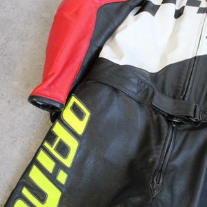 May include: Black and red leather motorcycle suit with white and black checkered accents. The suit features a yellow logo and lettering. The suit has a zipper closure and a belt.