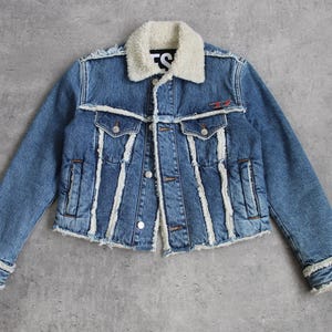 Puede incluir: Chaqueta vaquera azul con cuello y ribete de borreguillo color crema. La chaqueta tiene cierre de botones, dos bolsillos en el pecho y dos bolsillos laterales. Aspecto desgastado con bordes deshilachados y detalles blancos.