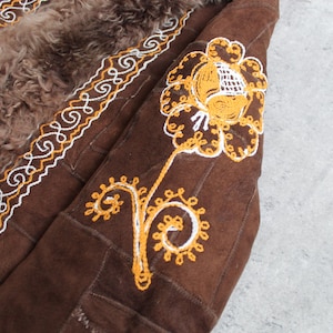 May include: Close-up of a brown suede coat with a sheepskin lining. The coat features intricate embroidery in white and yellow thread along the edges and on the sleeve, depicting a floral design. The coat has a vintage aesthetic.
