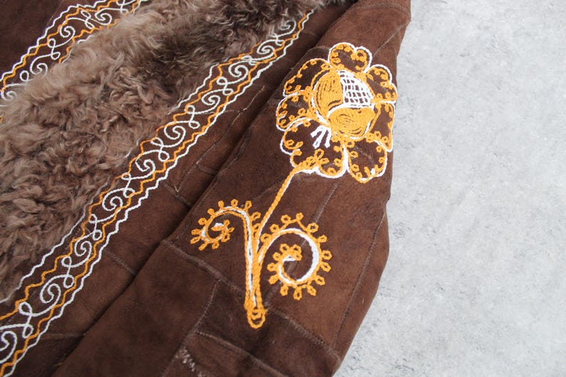 May include: Close-up of a brown suede coat with a sheepskin lining. The coat features intricate embroidery in white and yellow thread along the edges and on the sleeve, depicting a floral design. The coat has a vintage aesthetic.