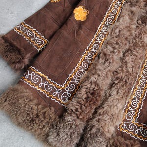 May include: Brown suede coat with intricate white and gold embroidered detailing. The coat features a plush, brown fur lining and trim. A yellow floral applique is visible on the upper portion of the coat. The coat is a vintage style.