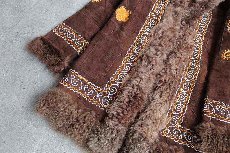 May include: Brown suede coat with intricate white and gold embroidered detailing. The coat features a plush, brown fur lining and trim. A yellow floral applique is visible on the upper portion of the coat. The coat is a vintage style.