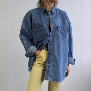 Pode incluir: Uma camisa de ganga azul com colarinho de botões e mangas compridas. A camisa tem dois bolsos no peito e é usada sobre calças amarelas.