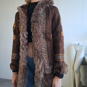 May include: A brown suede coat with a faux fur trim and hood. The coat features intricate embroidered detailing in white and gold along the front and cuffs. The coat has a vintage, bohemian style.