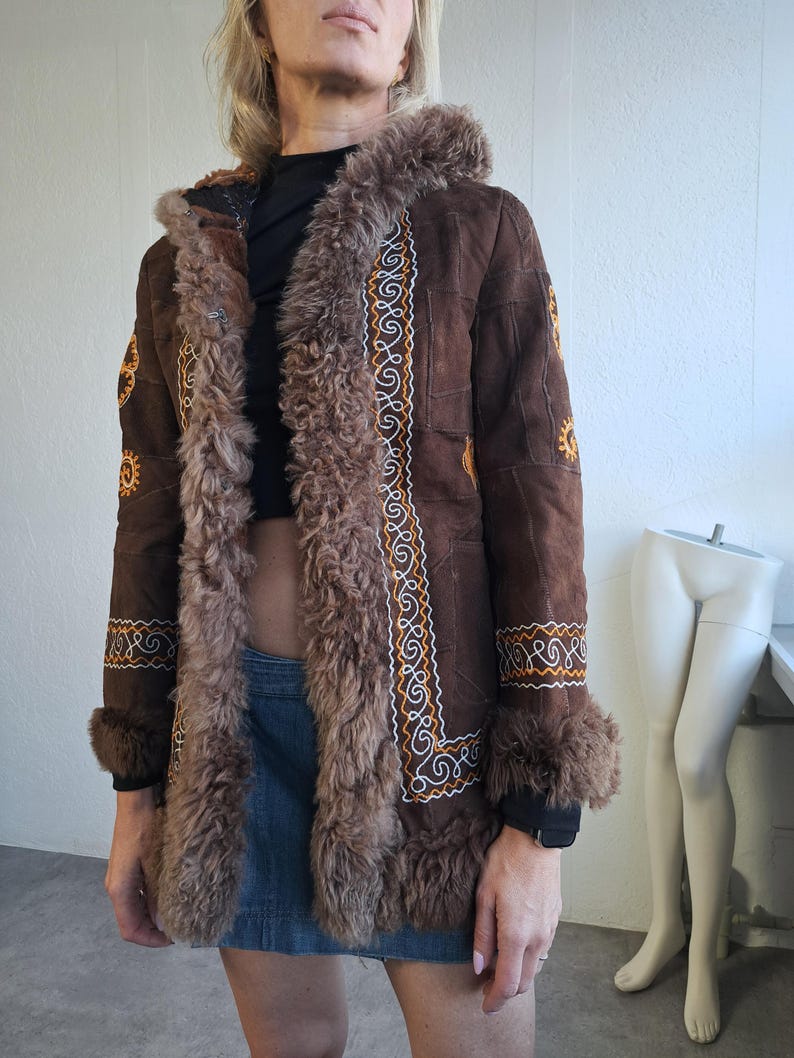 May include: A brown suede coat with a faux fur trim and hood. The coat features intricate embroidered detailing in white and gold along the front and cuffs. The coat has a vintage, bohemian style.
