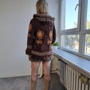 May include: A brown suede jacket with a hood and floral embroidery in orange and yellow. The jacket has fur trim around the hood, cuffs, and hem. The person is wearing tan knee-high boots.