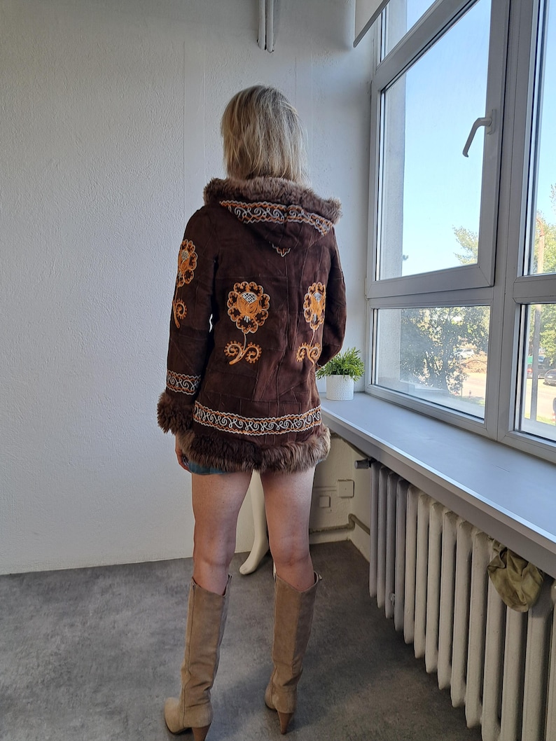 May include: A brown suede jacket with a hood and floral embroidery in orange and yellow. The jacket has fur trim around the hood, cuffs, and hem. The person is wearing tan knee-high boots.