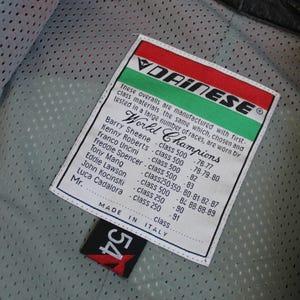 May include: Close-up of a black leather motorcycle jacket interior with a white tag. The tag features the Dainese logo, the words "World Champions," and a list of names and race years. A black tag with the number "54" is also visible.